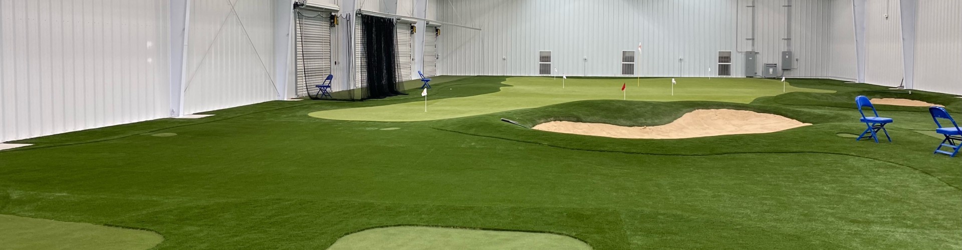 Southwest Greens indoor putting green facility with multiple holes, sand bunker, red and white flags inside a large commercial steel building.