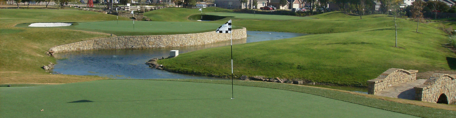 Artificial turf golf course with a checkered flag pin, stone retaining walls, water hazard, arched stone bridge, and multi-hole layout in a suburban residential setting