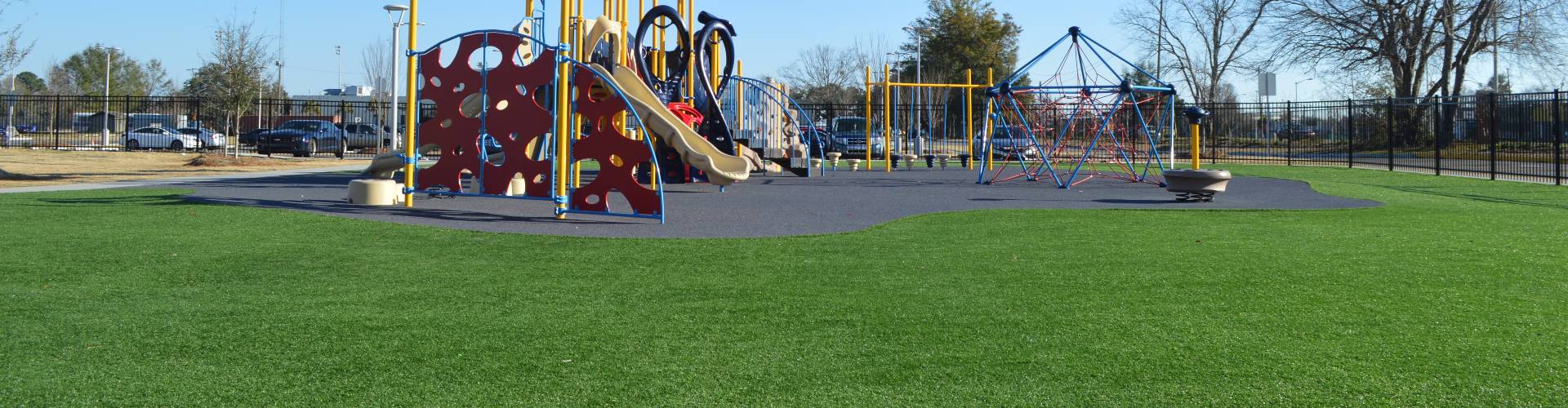 Fake grass installation surrounding a commercial playground with colorful climbing structures, slides, and rubber safety surfacing enclosed by a black iron fence