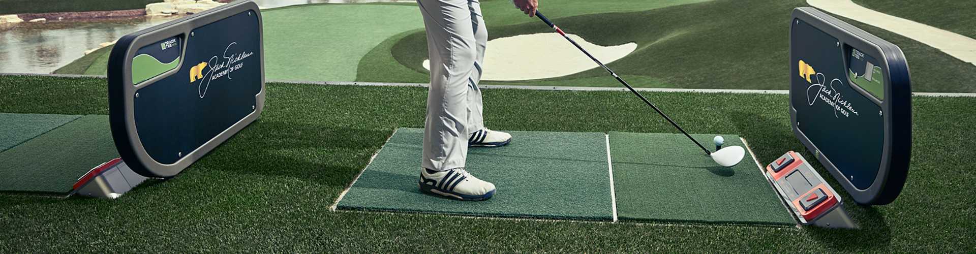 Golfer hitting an iron shot from an artificial turf golf course driving range bay with Jack Nicklaus Academy ball-tracking technology and a scenic course layout in the background