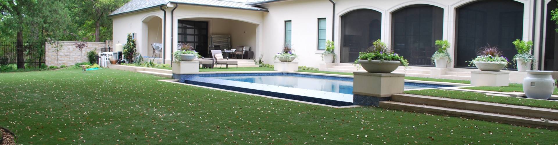 Best artificial lawns installed in a luxury residential backyard with a lap pool, covered patio, and mature oak trees surrounding a large stucco estate home